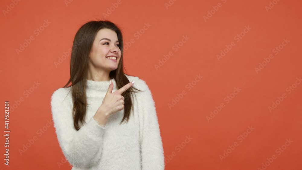 Look, advertise here! Smiling positive brunette woman in fluffy white sweater pointing finger away at empty background, freespace for advertisement. Indoor studio shot isolated on orange background Look, advertise here! Smiling positive brunette woman in fluffy white sweater pointing finger away at empty background, freespace for advertisement. Indoor studio shot isolated on orange background