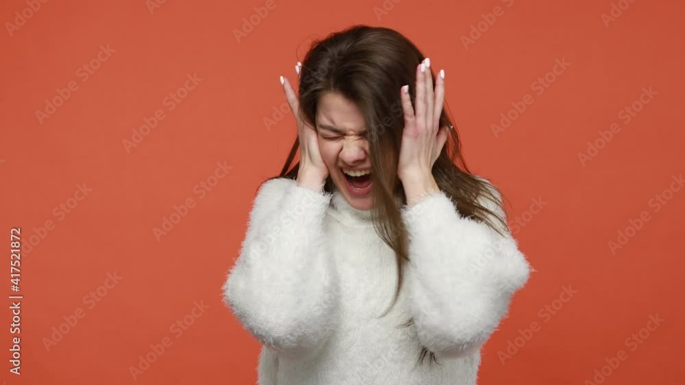 Scared panicked brunette woman in white fluffy sweater loudly screaming closing ears with hands, shocked and frightened, suffering paranoia. Indoor studio shot isolated on orange background Scared panicked brunette woman in white fluffy sweater loudly screaming closing ears with hands, shocked and frightened, suffering paranoia. Indoor studio shot isolated on orange background