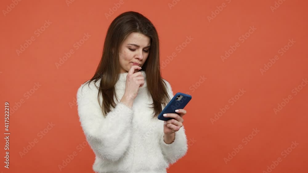 Concentrated pretty brunette woman in fluffy white sweater typing on smartphone, chatting in messenger or in social networks, commenting posts. Indoor studio shot isolated on orange background Concentrated pretty brunette woman in fluffy white sweater typing on smartphone, chatting in messenger or in social networks, commenting posts. Indoor studio shot isolated on orange background