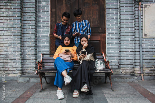 Four young friends in outdoors and looking at mobile phone. Two female sitting on a bench looking mobile phone with a frowny face and two male standing behind them smiling and holding smartphone