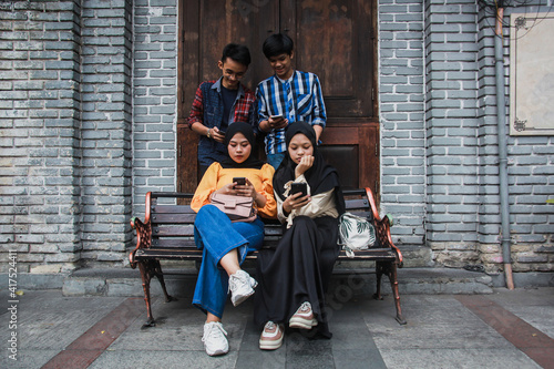 Four young friends in outdoors and looking at mobile phone. Two female sitting on a bench looking mobile phone with a frowny face and two male standing behind them smiling and holding smartphone photo