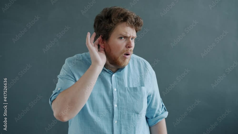 Slow motion portrait of guy touching ear trying to listen then spreading arms and looking at camera standing on gray background. People and body language concept. Slow motion portrait of guy touching ear trying to listen then spreading arms and looking at camera standing on gray background. People and body language concept.