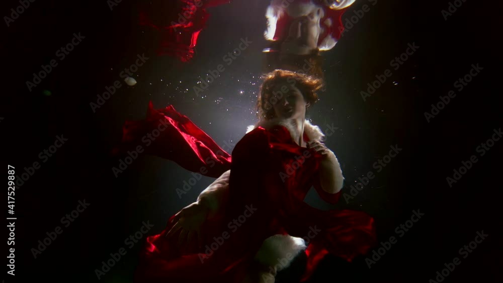 woman Santa underwater, young lady is dressed in red suit is floating in depth and playing with red silk cloth