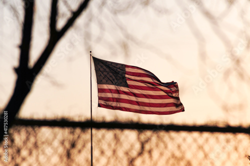 United States flag waving against sunset through tree branches and fence