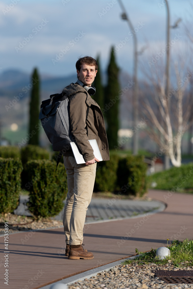 Full body portrait of a handsome student with a backpack and wearing winter clothes looking back to the camera outdoors in a park Full body portrait of a handsome student with a backpack and wearing winter clothes looking back to the camera outdoors in a park