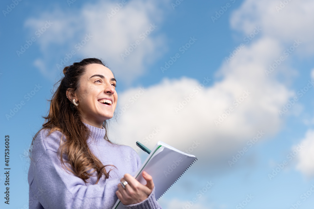 Closeup portrait of a smiling pretty woman writing on a notebook with a sky full of clouds as background. Closeup portrait of a smiling pretty woman writing on a notebook with a sky full of clouds as background.