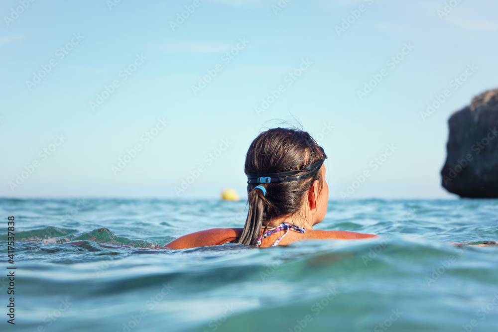 Young woman with swim goggles, surrounded by sea, view from behind, rocky cliff and clear sky background