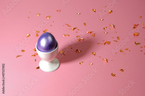 Easter egg of purple color in a white stand on a pink background with blurred sparkles. Background for your design. Congratulation or poster.