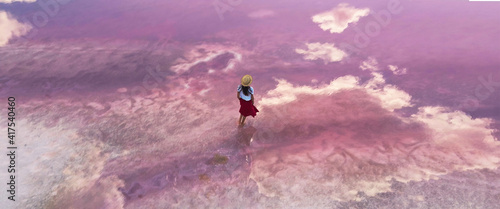 Aerial view girl in red skirt and straw hat walking in water of amazing pink lake with calm surface and clouds reflections.