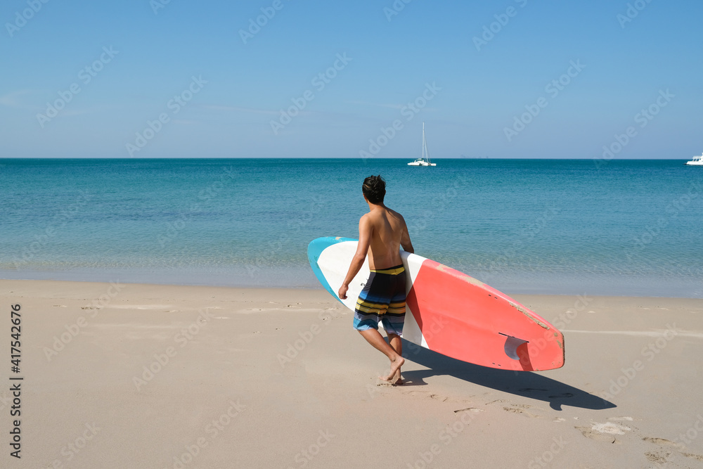 Obraz premium A sportman man with paddleboard going to the sea having fun and enjoying their vacations outdoors at the beach of Thailand. Surfer lifestyle. SUP or Stand Up Paddle Boarding.