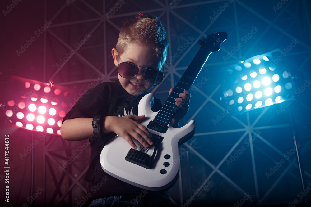 Obraz premium Little boy in sunglasses with a guitar on a background of colored lights. The inscription on the T-shirt Rock music. Hobby and leisure concept.