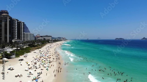 Drone flying downwards in Barra da Tijuca beach while waves crash on beach. morning traffic near noon, lots of people on the beach on beautiful sunny day