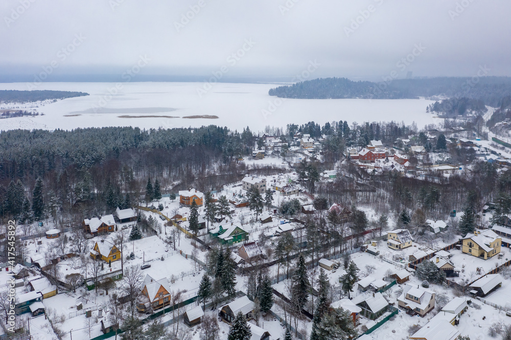 Aerial view of a snow-covered village near the lake. Winter rural landscape. Top view of houses and trees. Cold snowy winter weather. Snowfall. Snow on the roofs. Toksovo, Leningrad region, Russia.