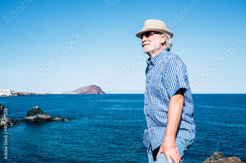 Portrait of smiling senior man with hat in front to the sea. Blue sky and mountain in the distance. Horizon over water