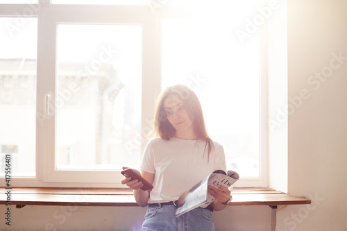 Beautiful millenial woman using mobile phone and smiling while standing near window. Natural morning light. Chatting or shopping online