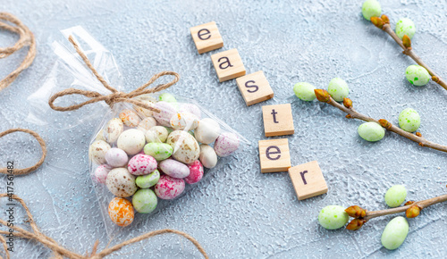 Chocolate Easter eggs in glaze in a transparent bag, branches of a spring tree and wooden letters on a light blue background. Happy Easter time. Preparation for holiday, gift wrapping. Banner.