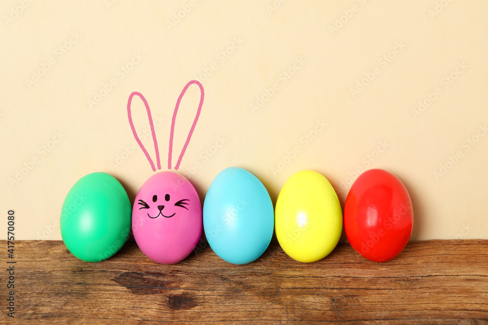 One eggs with drawn face and ears as Easter bunny among others on wooden table against beige background