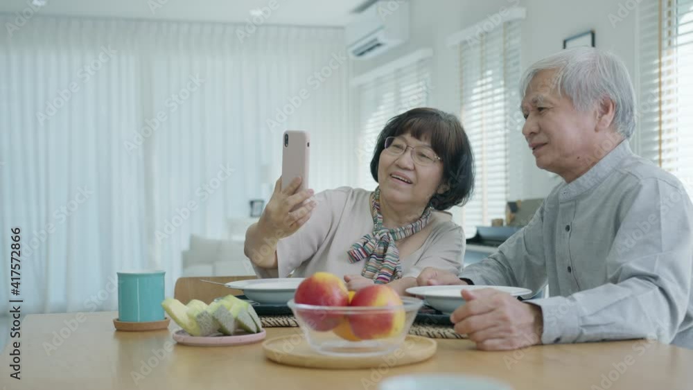 Young senior asia citizen couple hold mobile phone selfie videocall online to family at home apartment morning on dining table in older people with digital technology virtual talk conference concept. Young senior asia citizen couple hold mobile phone selfie videocall online to family at home apartment morning on dining table in older people with digital technology virtual talk conference concept.
