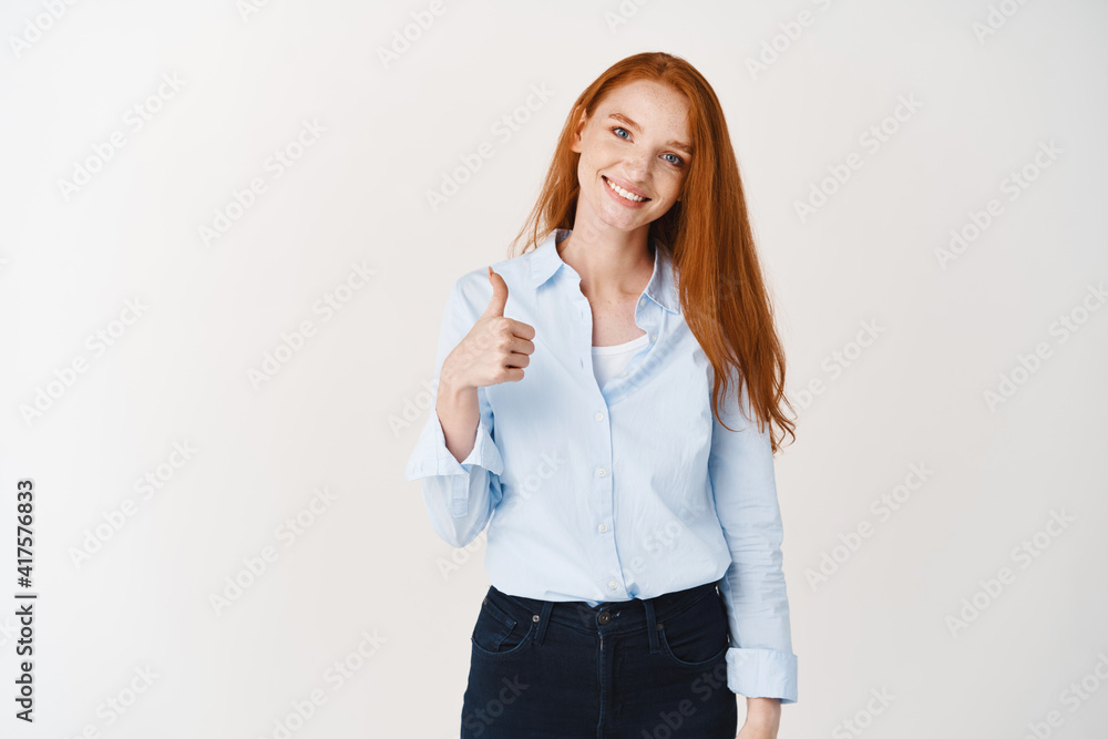 Young smiling redhead woman in blue shirt showing thumbs-up, praising good job, approve and agree, standing over white background Young smiling redhead woman in blue shirt showing thumbs-up, praising good job, approve and agree, standing over white background