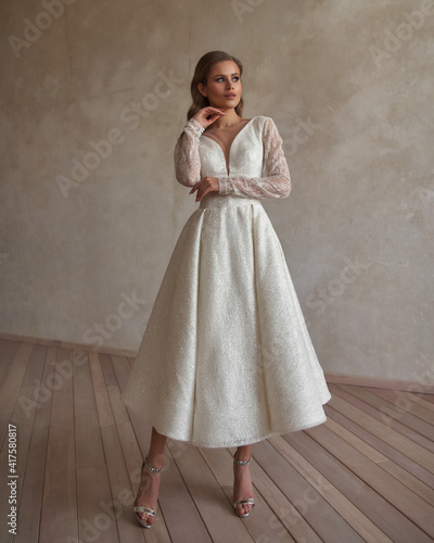 Full length portrait of young beautiful woman wearing white wedding dress. Elegant bride standing and posing against beige wall