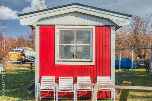 A typical Swedish red summerhouse with a square window and white chairs. The bare exterior has a simple aspect. Its wooden facade and the boats in the yard suggest it belongs to a local fisherman photo