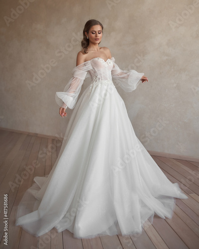 Full length bride portrait. Elegant woman in white wedding dress spinning in beige interior. Dynamic shot. Elegant female model standing and posing in ball gown