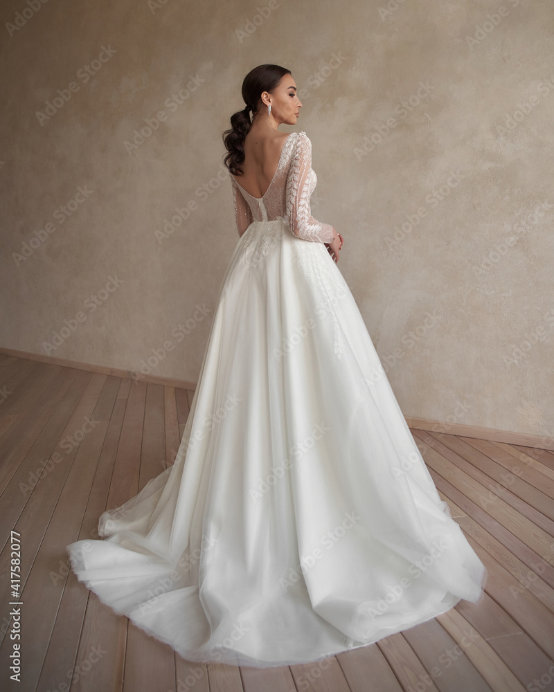 Full length portrait of young beautiful woman wearing white wedding dress. Elegant bride standing and posing against beige wall Full length portrait of young beautiful woman wearing white wedding dress. Elegant bride standing and posing against beige wall