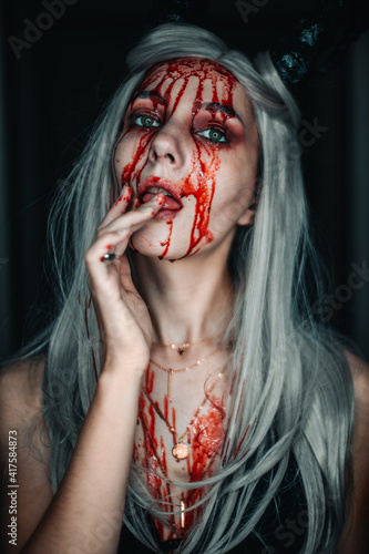 A beautiful demon girl with blood running down her face licks the blood from her hand. On the head of the horns, on the face of a beautiful makeup