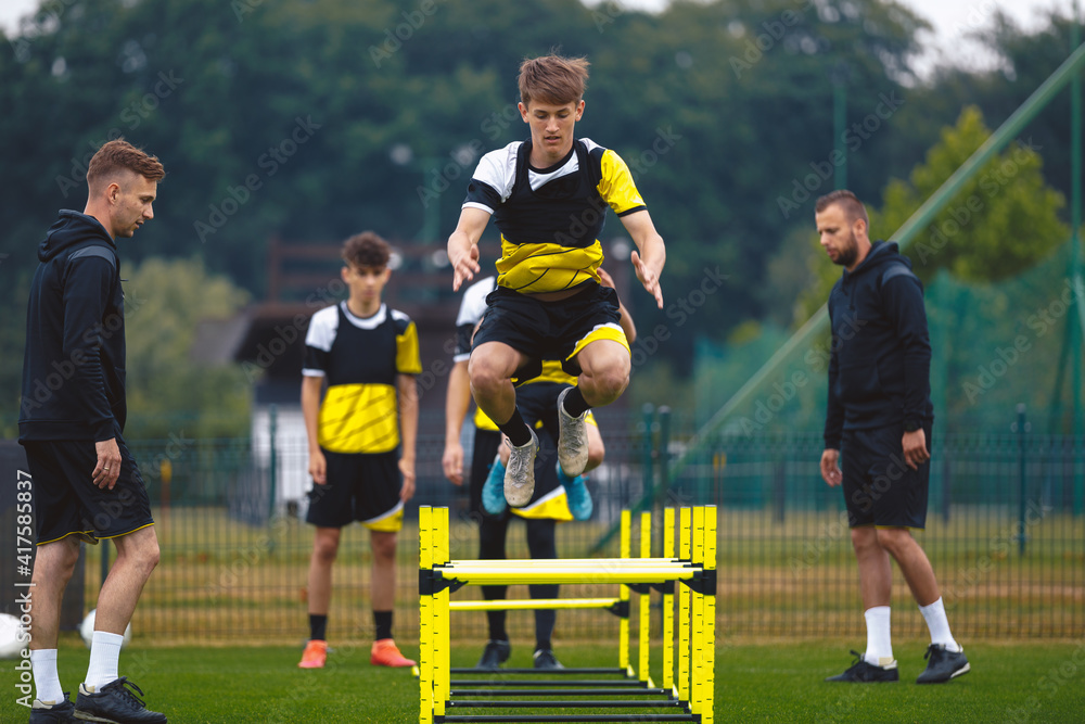 Obraz premium Youth Soccer Team on Training. Young Boys in Football Club Practicing Durability. Teenage Boy Jumping High Over Hurdle Obstacles. Young Men Coaching Sports Team on Practice Field. Youth Soccer School