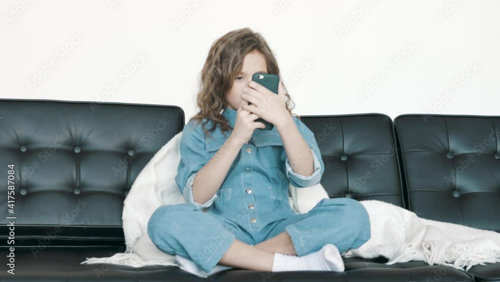 Cute kid girl holding smart phone enjoying using mobile apps, playing games at home. Small mixed race child learning in cellphone, watching video, having fun with mobile technology concept.