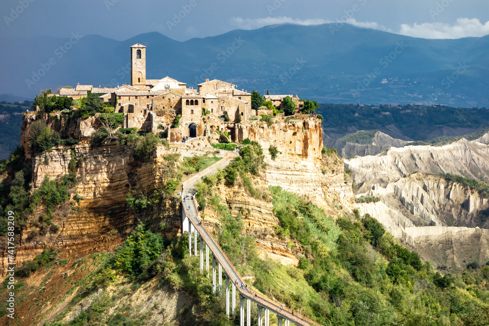 View of sunlit village of Bagnoregio, Italy, with dark mountains and clouds in the background