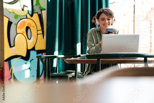 Girl in headphones using cellphone and laptop while sitting in cafe