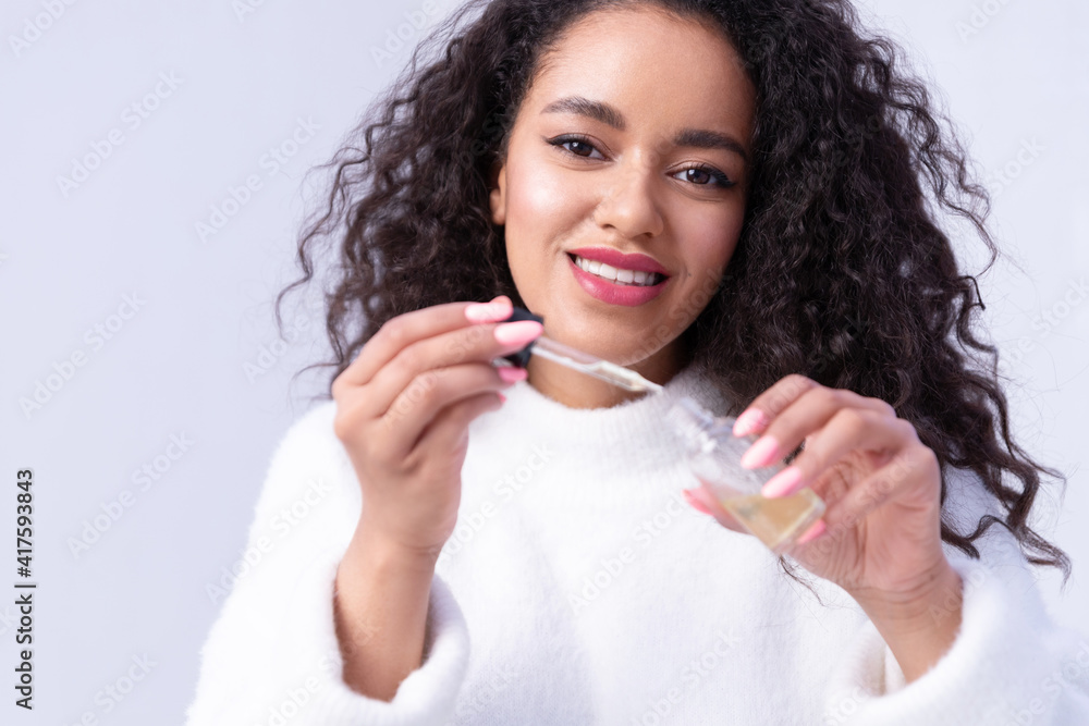 Smiling young black girl holding bottle and pipette of hypoallergenic delicate serum or natural organic oil. Positive mixed race girl show jar of skincare moisturizing nourishing cosmetic product Smiling young black girl holding bottle and pipette of hypoallergenic delicate serum or natural organic oil. Positive mixed race girl show jar of skincare moisturizing nourishing cosmetic product