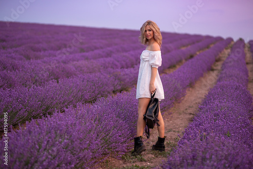 Side view of la blonde young lady in delicate white dress and black boots looking at camera while sitanding alone among aromatic lavender flowers on meadow photo