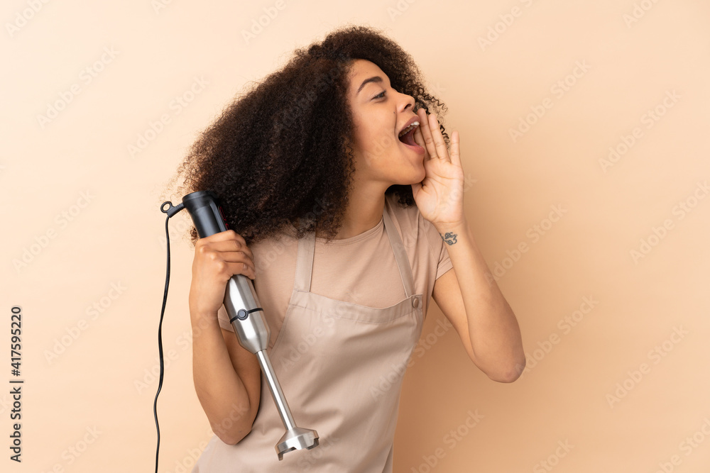 Young african american woman using hand blender isolated on beige background shouting with mouth wide open Young african american woman using hand blender isolated on beige background shouting with mouth wide open