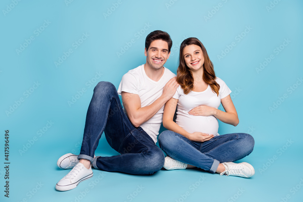 Photo of optimistic pregnant couple sit hug wear white t-shirts jeans sneakers isolated on cyan color background Photo of optimistic pregnant couple sit hug wear white t-shirts jeans sneakers isolated on cyan color background