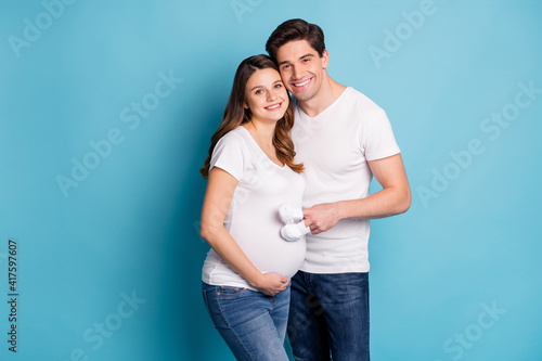 Photo of positive young spouse hugging toothy smile baby booties on fingers step stomach isolated on blue color background