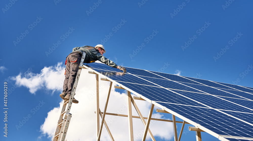 Side view of male worker standing on ladder and installing blue photovoltaic solar panel under beautiful cloudy sky. Concept of sustainable energy and ecological solution.