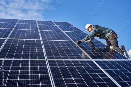 Horizontal snapshot of beautiful and shiny solar battery surface and male worker wearing a uniform, standing on ladder, installing solar modules on sunny day, low angle view