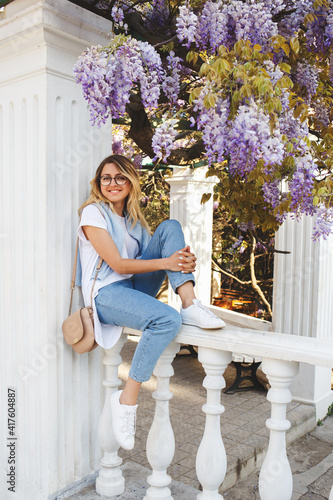 Young beautiful woman in jeans, white t-shirt, sneakers and glasses sitting near a flowering Wisteria tree. Spring portrait.