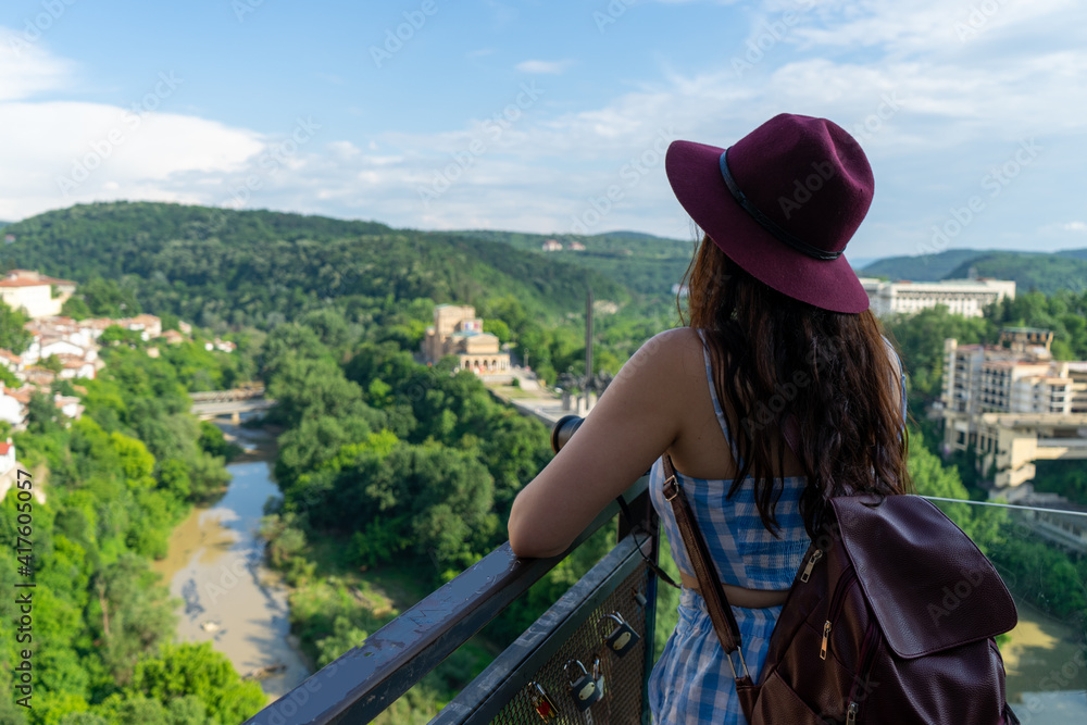 White pretty female wear hat, enjoying the vista in a open balcony. Backpacker in blue summer dress and bag doing a trip in the balkans. Contemplation of panoramic in a tour.