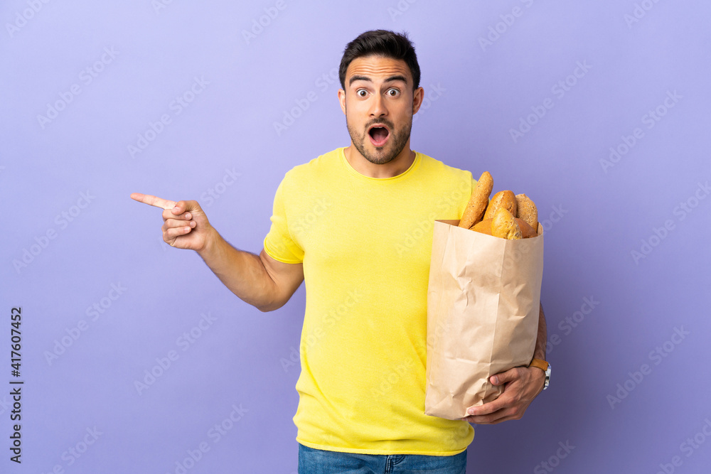 Young caucasian man holding a bag full of breads isolated on purple background surprised and pointing finger to the side Young caucasian man holding a bag full of breads isolated on purple background surprised and pointing finger to the side