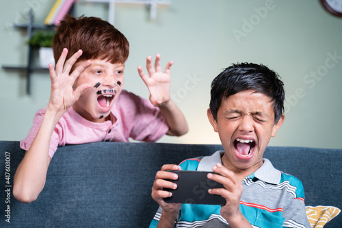 Fotografi young kid with face painting fools or making prank to his brother while he is busy on mobile by suddenly screaming and shouting from back side at home during april fools day celebration