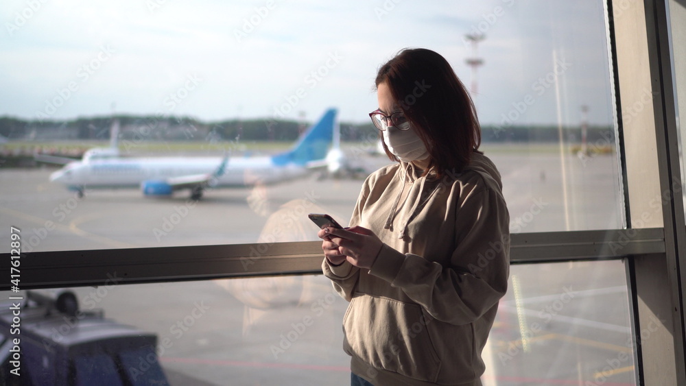 Young woman in a medical mask with a phone in her hands on the background of a window at the airport. Airplanes in the background.