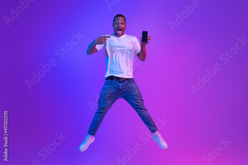 Amazed African Man Jumping Over Purple Neon Background And Pointing At Smartphone