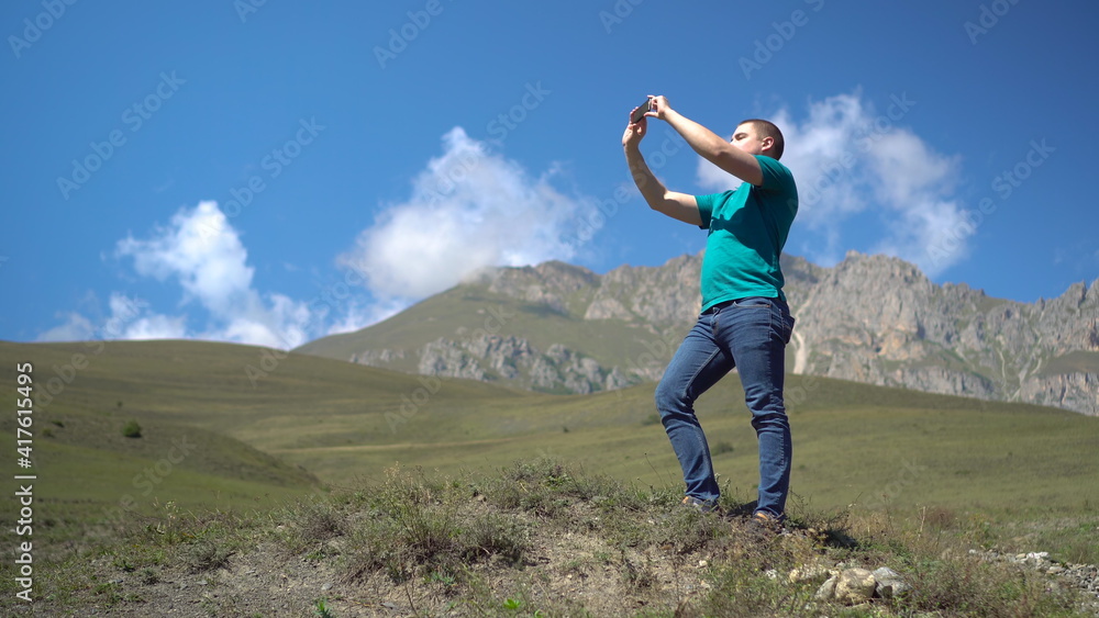 A young man stands in the mountains and takes pictures on the phone. A man travels in the Caucasian mountains.