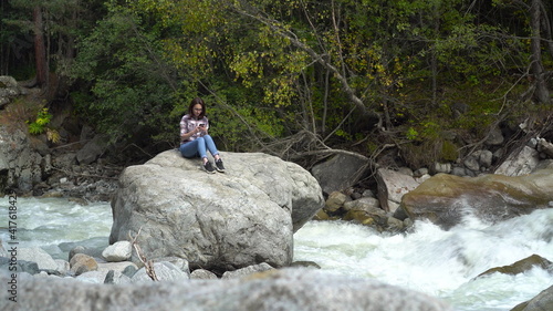 A young girl sits on a stone with a phone in her hands in the mountains by the river. Travel to the mountains.