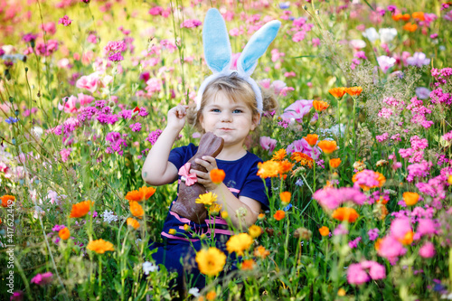Portriat of adorable, charming toddler girl with Easter bunny ears eating chocolate bunny figure in flowers meadow. Smiling happy baby child on sunny day with colorful flowers, outdoors. © Irina Schmidt