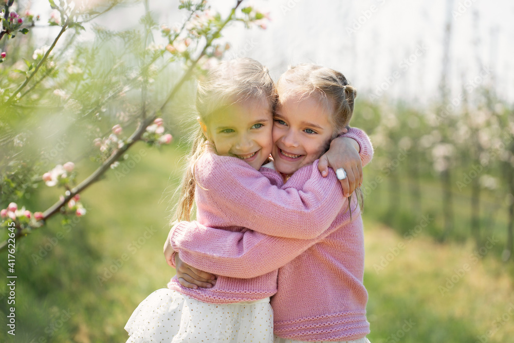 Happy two twin sisters are hugging against the background of a green blossoming apple orchard Happy two twin sisters are hugging against the background of a green blossoming apple orchard