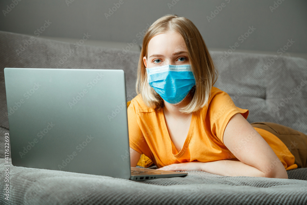 Teen girl in mask do Online learning education via laptop covid 19 lockdown time. Remote work in Coronavirus pandemic. Woman in medical protective mask work using laptop at Home office lies on couch. Teen girl in mask do Online learning education via laptop covid 19 lockdown time. Remote work in Coronavirus pandemic. Woman in medical protective mask work using laptop at Home office lies on couch.